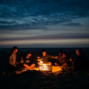Kompisgäng sitter runt Höfats Cube Eldkorg & Kolgrill rost på stranden på kvällen