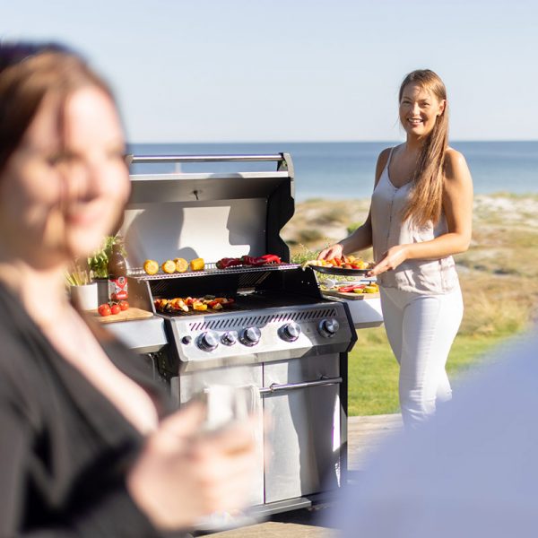 Kvinna håller tallrik med grillad mat bredvid Bluegaz X1 Premium Classic Gasolgrill med havet i bakgrund