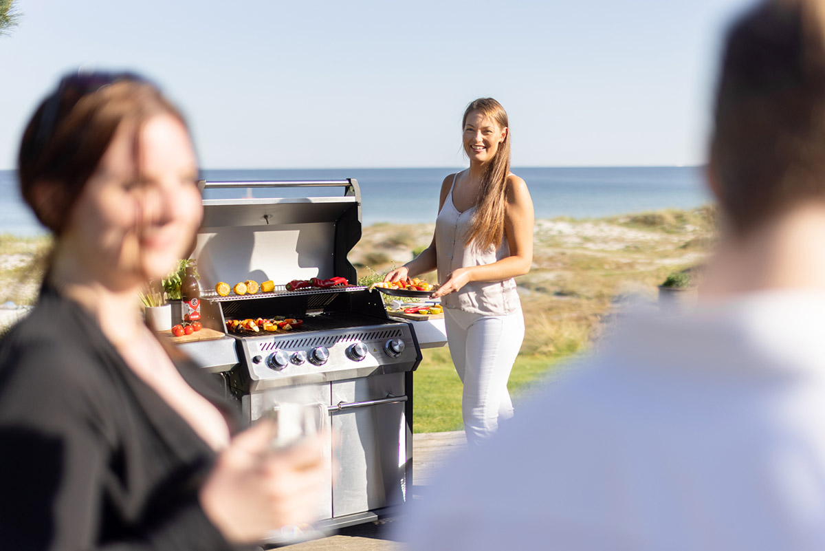 Kvinna håller tallrik med grillad mat bredvid Bluegaz X1 Premium Classic Gasolgrill med havet i bakgrund