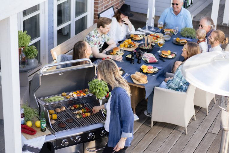 Tjej grillar hamburgare och grillspett på Bluegaz Z1 Gasolgrill med sällskap som sitter vid bord bredvid