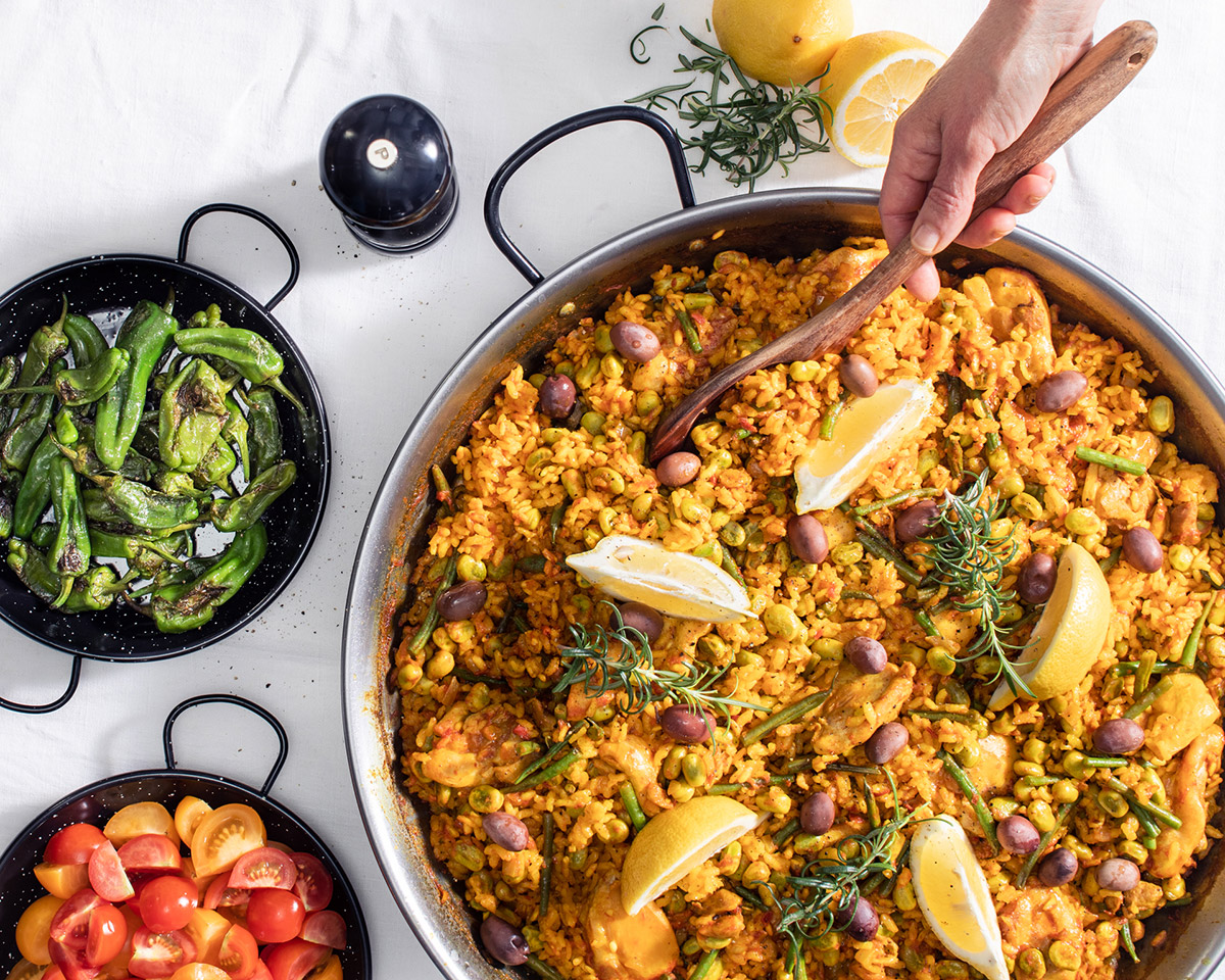 Hand håller i träslev för att servera Paella Valenciana med kyckling direkt från paellapannan