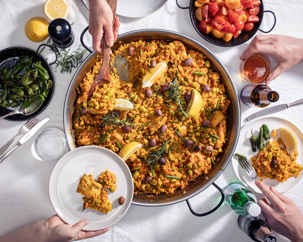 Hand håller i träslev för att servera sällskap och tar upp Paella Valenciana med kyckling direkt från paellapannan som står på ett matbord med andra läckerheter vid sidan av