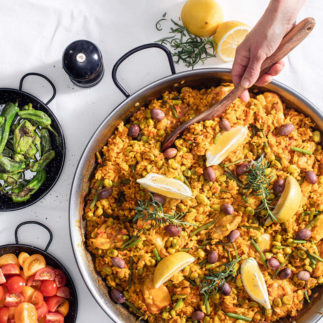 Hand håller i träslev för att servera Paella Valenciana med kyckling direkt från paellapannan