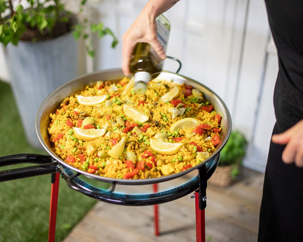 Kvinna häller i olivolja i vegetarisk paella med halloumi, svamp och kronärtskockor som tillagas i Bluegaz paellaset