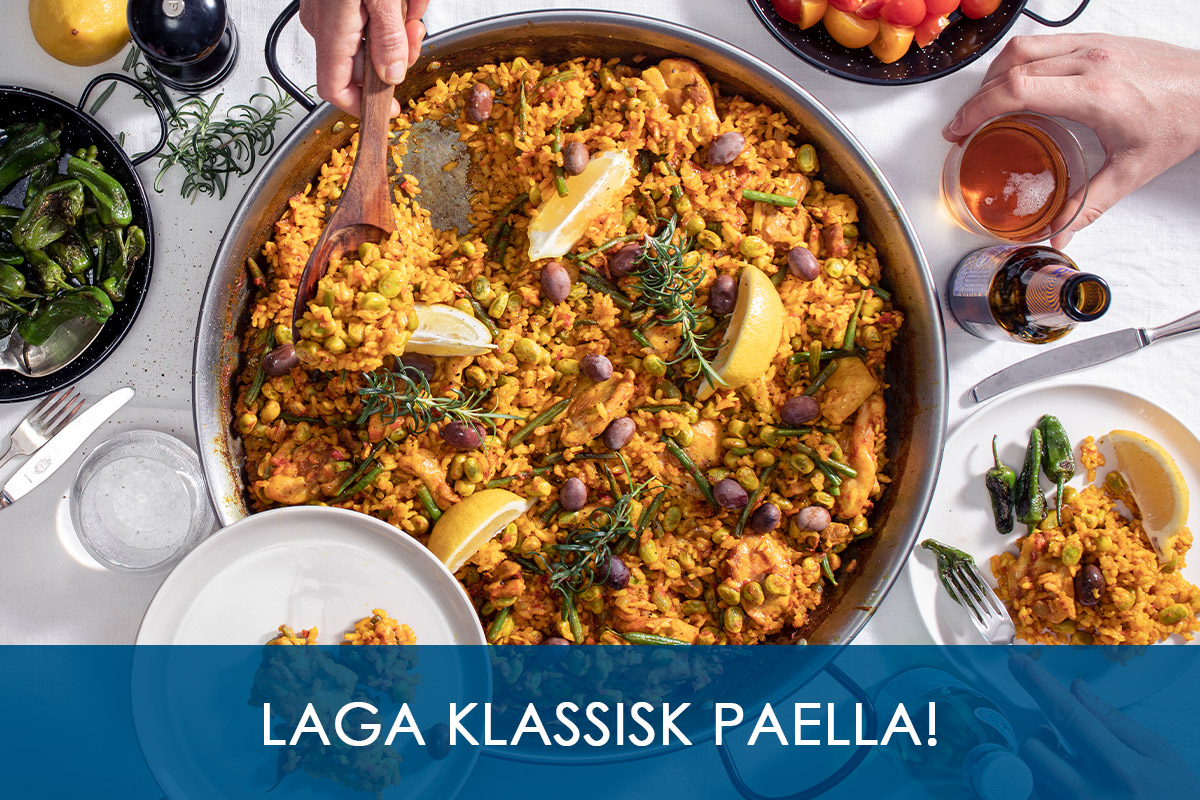 Hand håller i träslev för att servera sällskap och tar upp Paella Valenciana med kyckling direkt från paellapannan som står på ett matbord med andra läckerheter vid sidan av