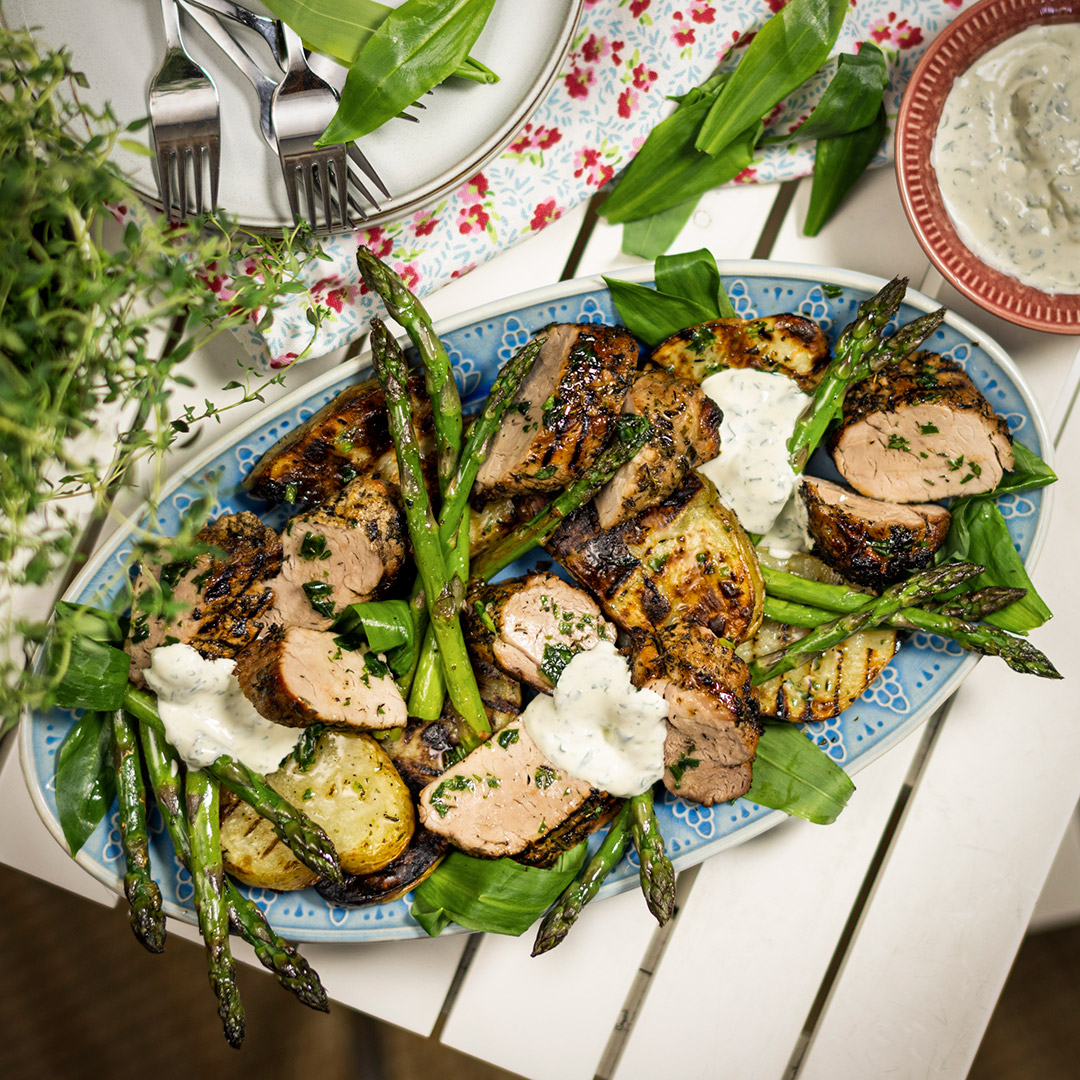 Blå tallrik med grillad fläskfilé, sparris och skivad bakpotatis på vitt bord samt röd liten skål med ramslökssås