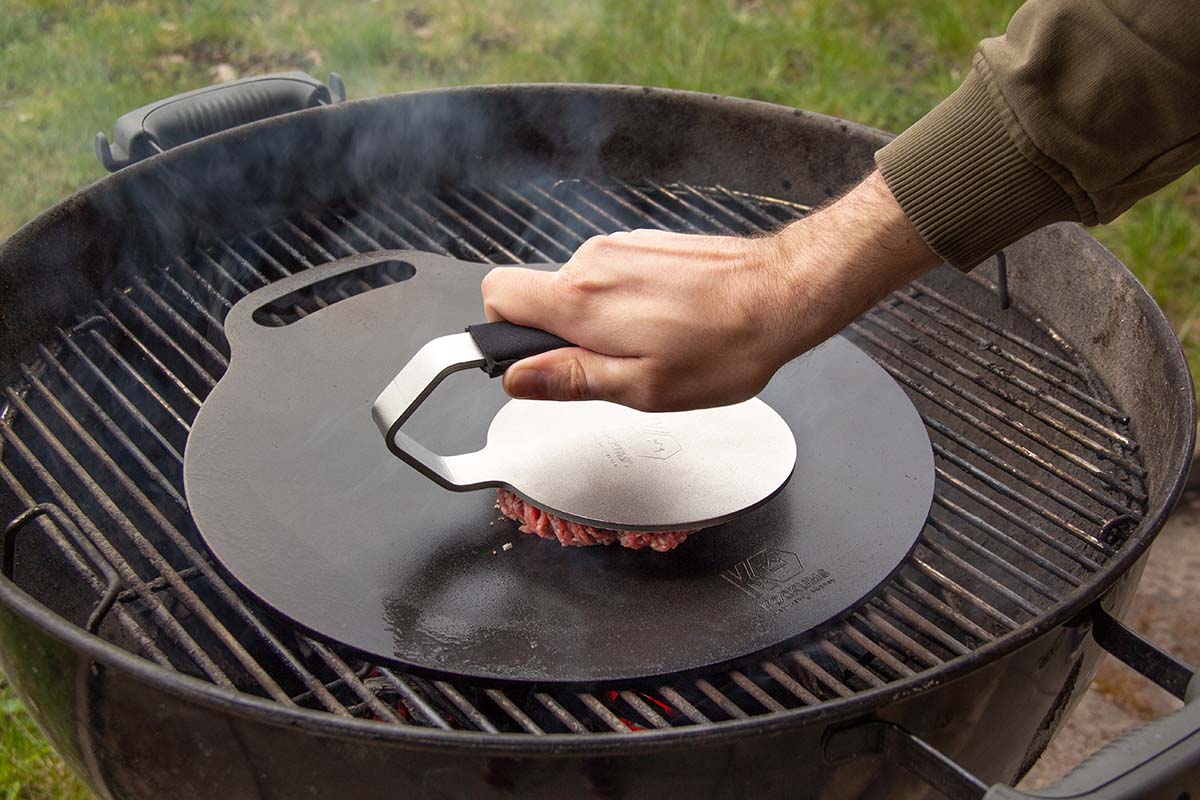 Man smashar hamburgare med Kockums Jernverk smashjärn med neoprenskydd på stekplatta på kolgrill