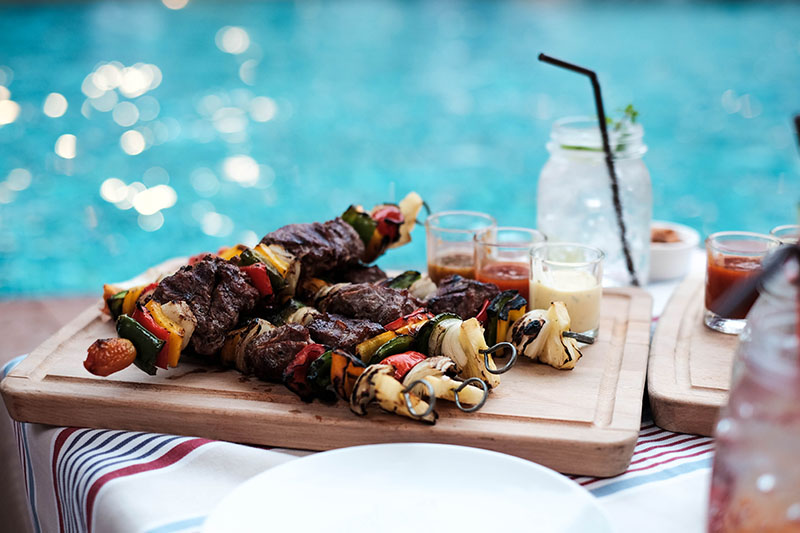 Ljus skärbräda med grillspett bestående av kött, paprika, lök och tre olika dressings framför pool