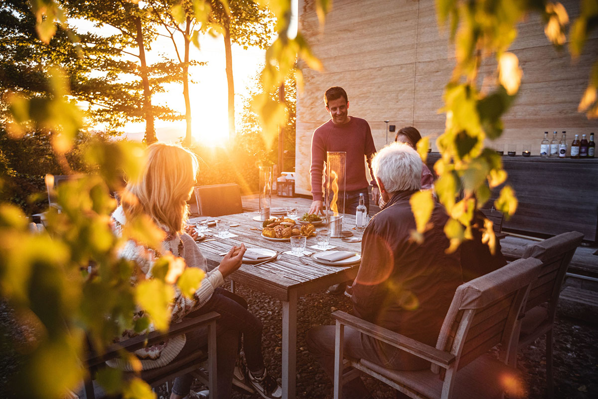 Familj sitter vid bord utomhus och äter middag framför SPIN Eldlykta