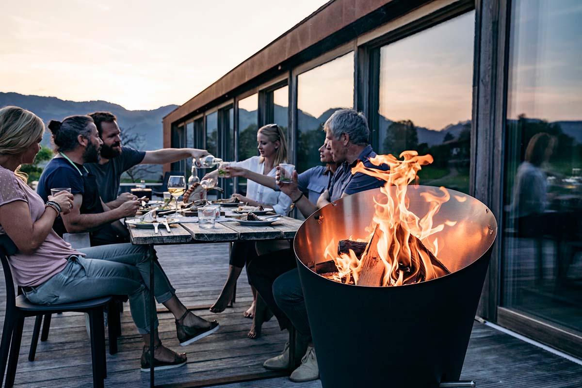 Ved eldas i Höfats Cone Kolgrill med sällskap som äter grillmiddag i bakgrunden under kvällstid