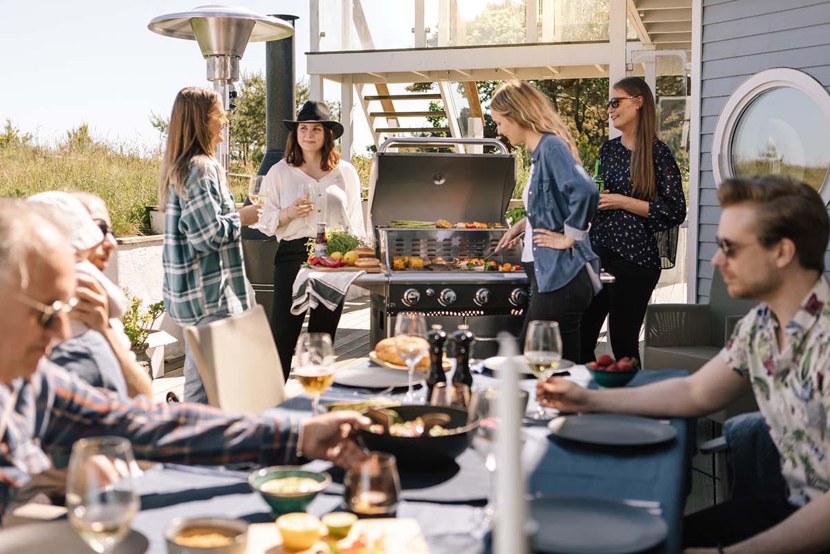 Tjejgäng grillar på Bluegaz Z1 Gasolgrill med sällskap vid bord i bakgrunden