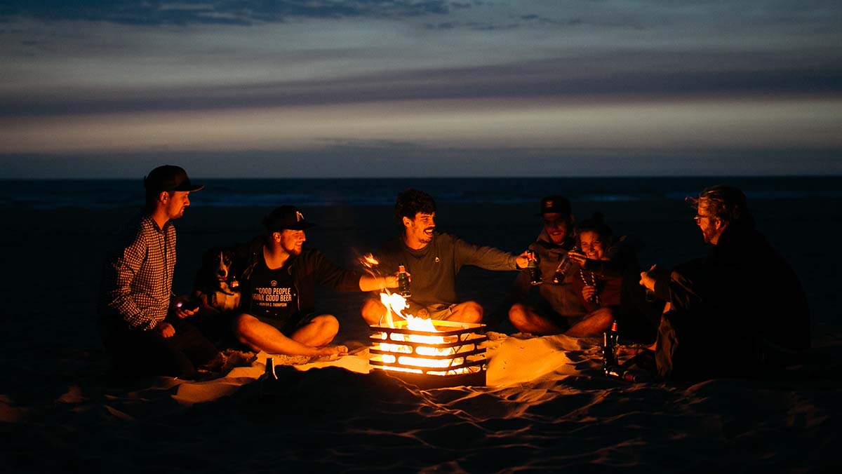 Kompisgäng sitter runt Höfats Cube Eldkorg & Kolgrill rost på stranden på kvällen