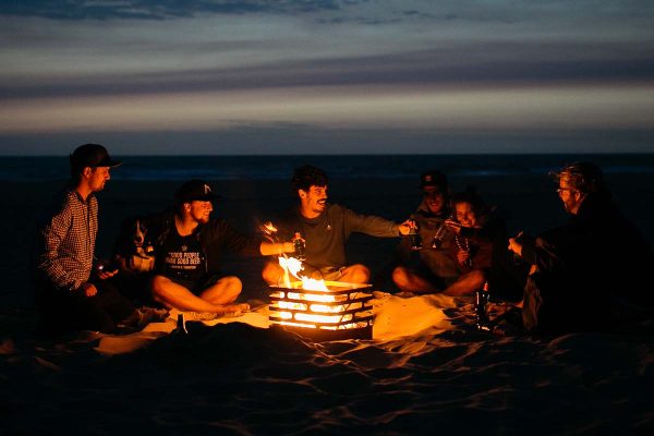 Kompisgäng sitter runt Höfats Cube Eldkorg & Kolgrill rost på stranden på kvällen