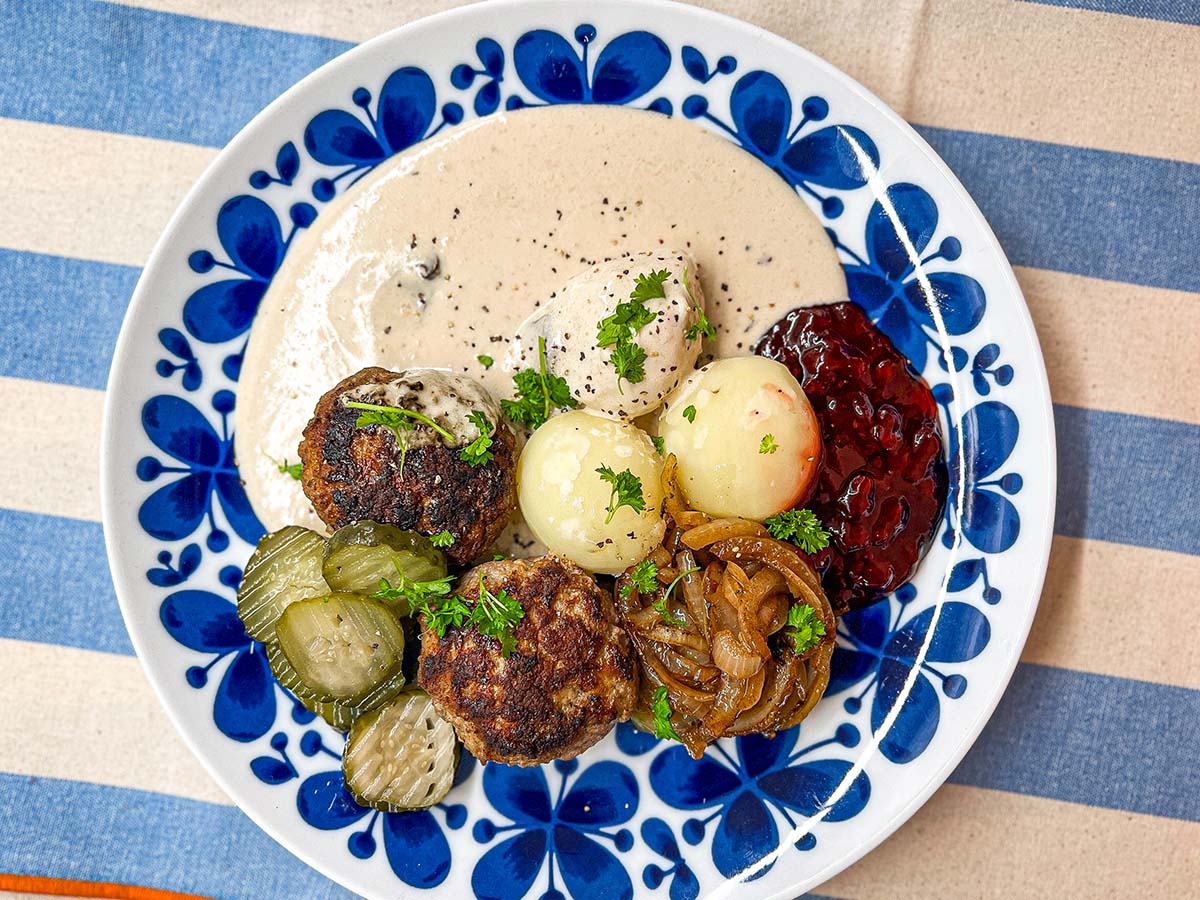 Pannbiff, potatis, gul lök, saltgurka, lingonsylt och gräddsås på vit och blå tallrik på duk