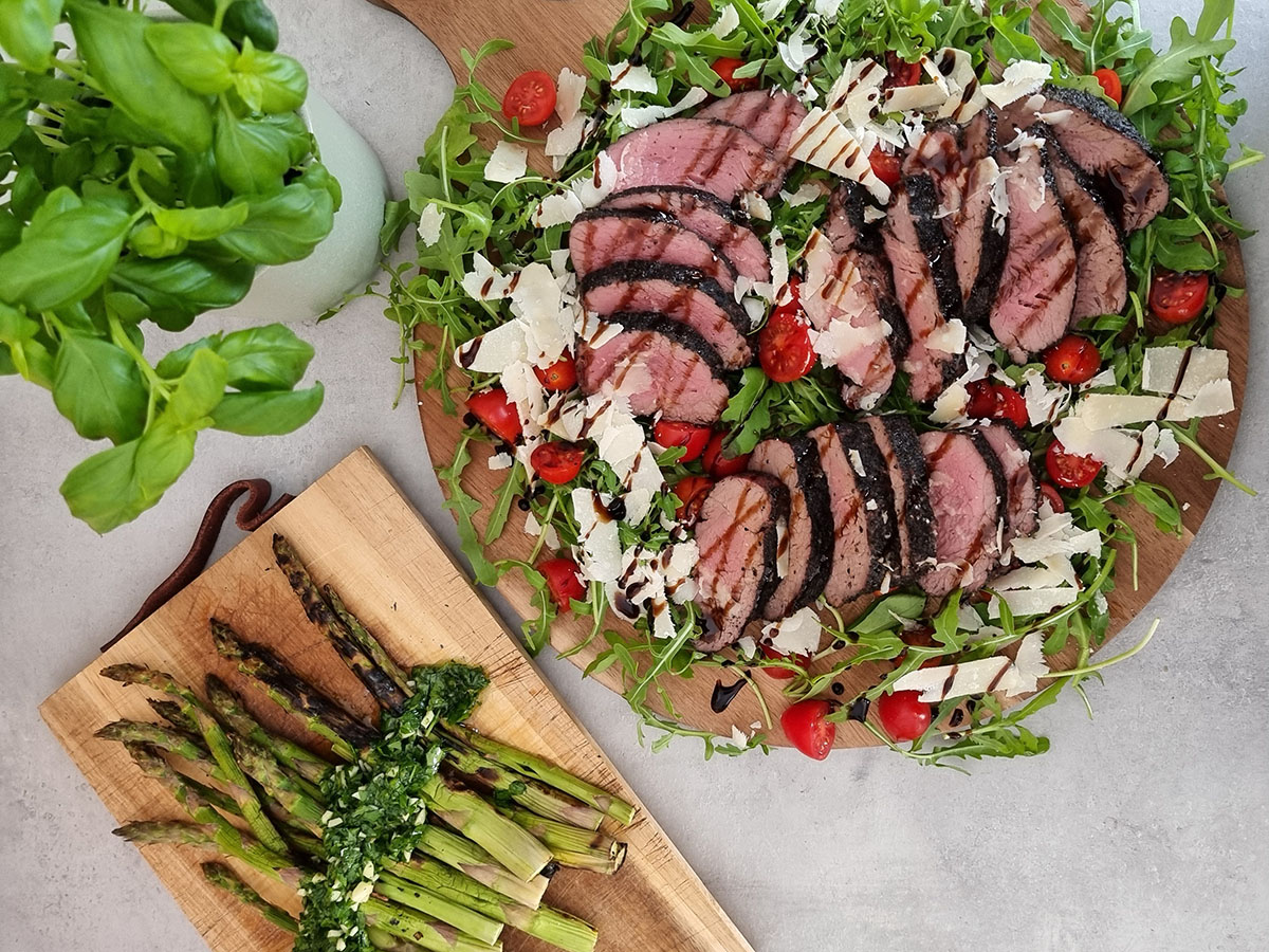 Grillad tagliata, ruccola, parmesan och körsbärstomater på rund skärbricka bredvid skärbräda med grillad sparris och silverlök