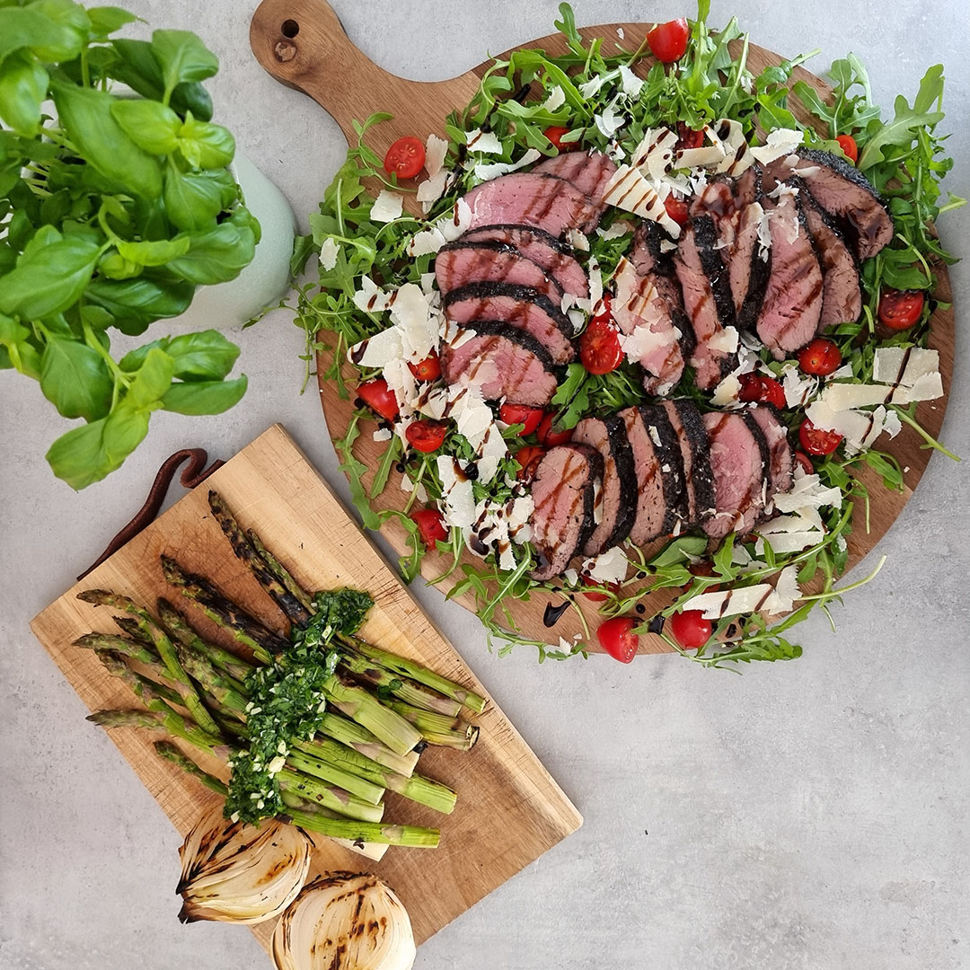 Grillad tagliata, ruccola, parmesan och körsbärstomater på rund skärbricka bredvid skärbräda med grillad sparris och silverlök