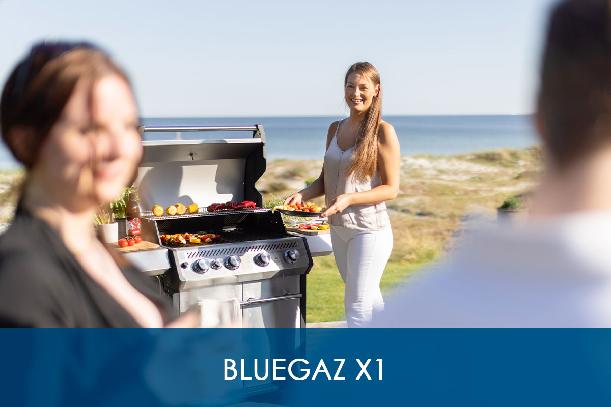 Kvinna håller tallrik med grillad mat bredvid Bluegaz X1 Premium Classic Gasolgrill med havet i bakgrund