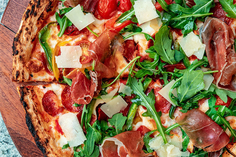 Pizza med prosciutto, ruccola och parmesan på rund skärbräda i mörkt trä