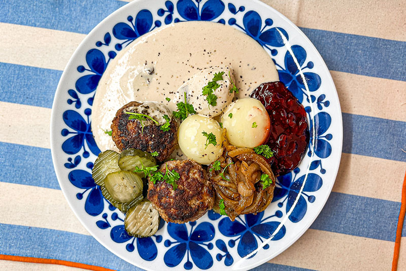 Pannbiff, potatis, gul lök, saltgurka, lingonsylt och gräddsås på vit och blå tallrik på duk