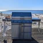 Bluegaz Z501 Gasolgrill rostfri står på trädäck med strand och hav i bakgrunden