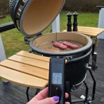 Håller i Bluegaz trådlös grilltermometer med 2 sonder framför kamadogrill med två köttbitar i